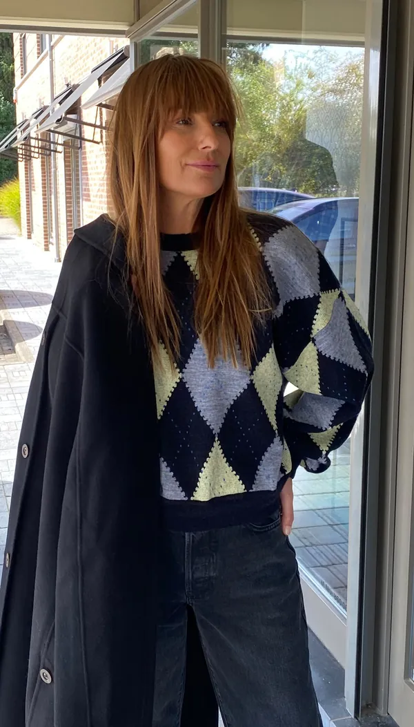 Collection Hiver 2026 : pull à losanges marine et anis, manteau noir sur l'épaule